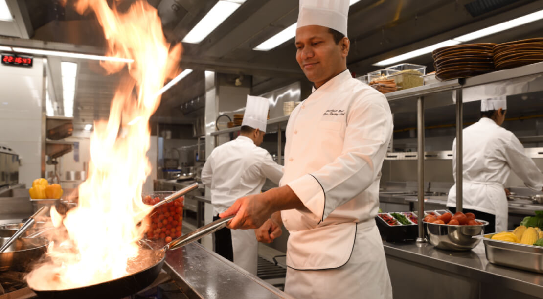 A chef in a white uniform and hat cooks with flames leaping from a pan in a busy, stainless steel kitchen
