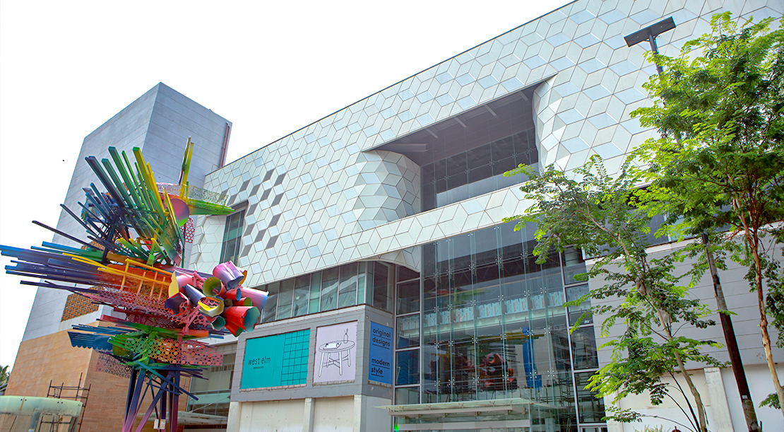A modern building with a geometric facade and large glass windows. In front, theres a colorful abstract sculpture made of various materials and shapes. Trees partially frame the scene, and a digital signboard is visible on the building